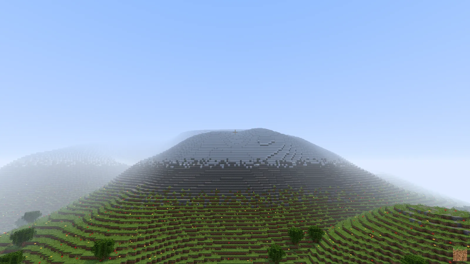 A foggy grey stone mountain rising above dark grass terraces under a pale blue sky